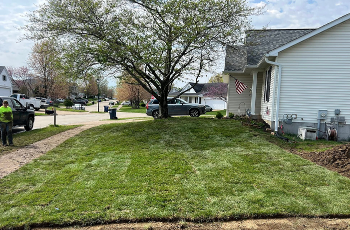 Freshly laid sod on a suburban front lawn, bordered by a driveway and white house. A flag hangs near the house, and a person stands beside a parked truck.