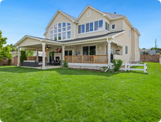 A spacious beige two-story house with large windows and a covered patio, surrounded by a lush green lawn under a clear, bright blue sky.