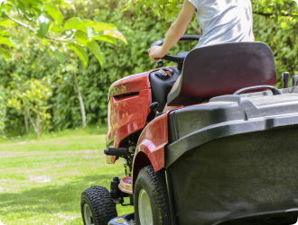 A person rides a red lawn tractor across a green yard, bordered by lush trees, conveying a sense of outdoor activity and maintenance.