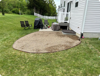A backyard patio with gravel, adjacent to a white house. Patio furniture and a grill are arranged on the paved area. Green lawn and trees surround the space.