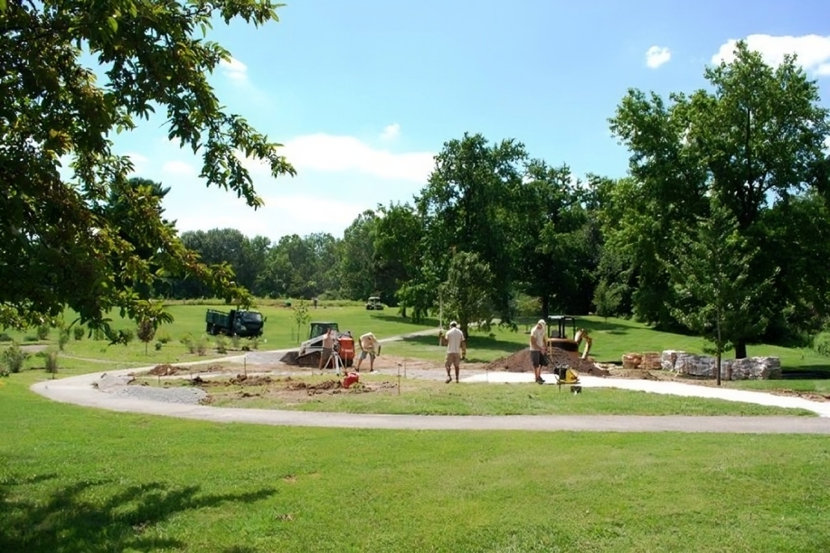 Workers landscaping a park under a sunny sky, surrounded by green grass and tall trees. Construction materials and equipment are visible nearby.