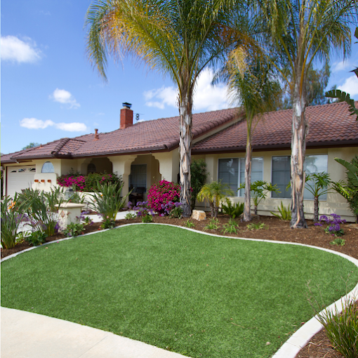 Single-story house with a red-tiled roof and tan walls, surrounded by lush greenery and palm trees, featuring a manicured lawn and colorful flowers.