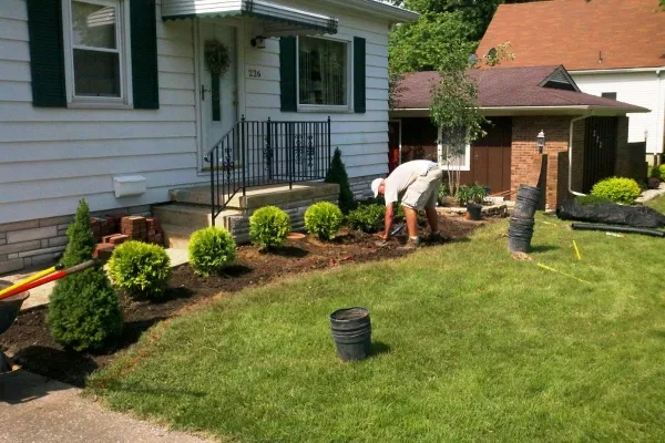 A person in a white shirt tends to a garden in front of a house with white siding and dark shutters. Green shrubs line the garden bed, and a garage is visible in the background. The scene is sunny and peaceful, conveying a sense of care and home improvement.