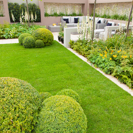 A lush garden with manicured green grass, rounded bushes, vibrant flowers, and a cozy gray patio area featuring cushioned seating. Tranquil ambiance.